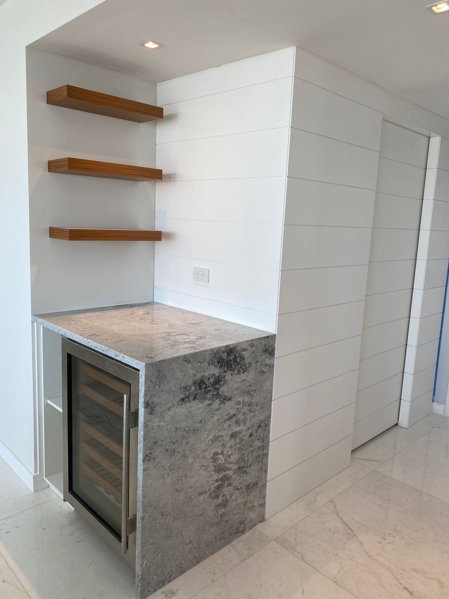 Modern kitchen corner with floating wooden shelves and a small built-in refrigerator under a marble countertop.