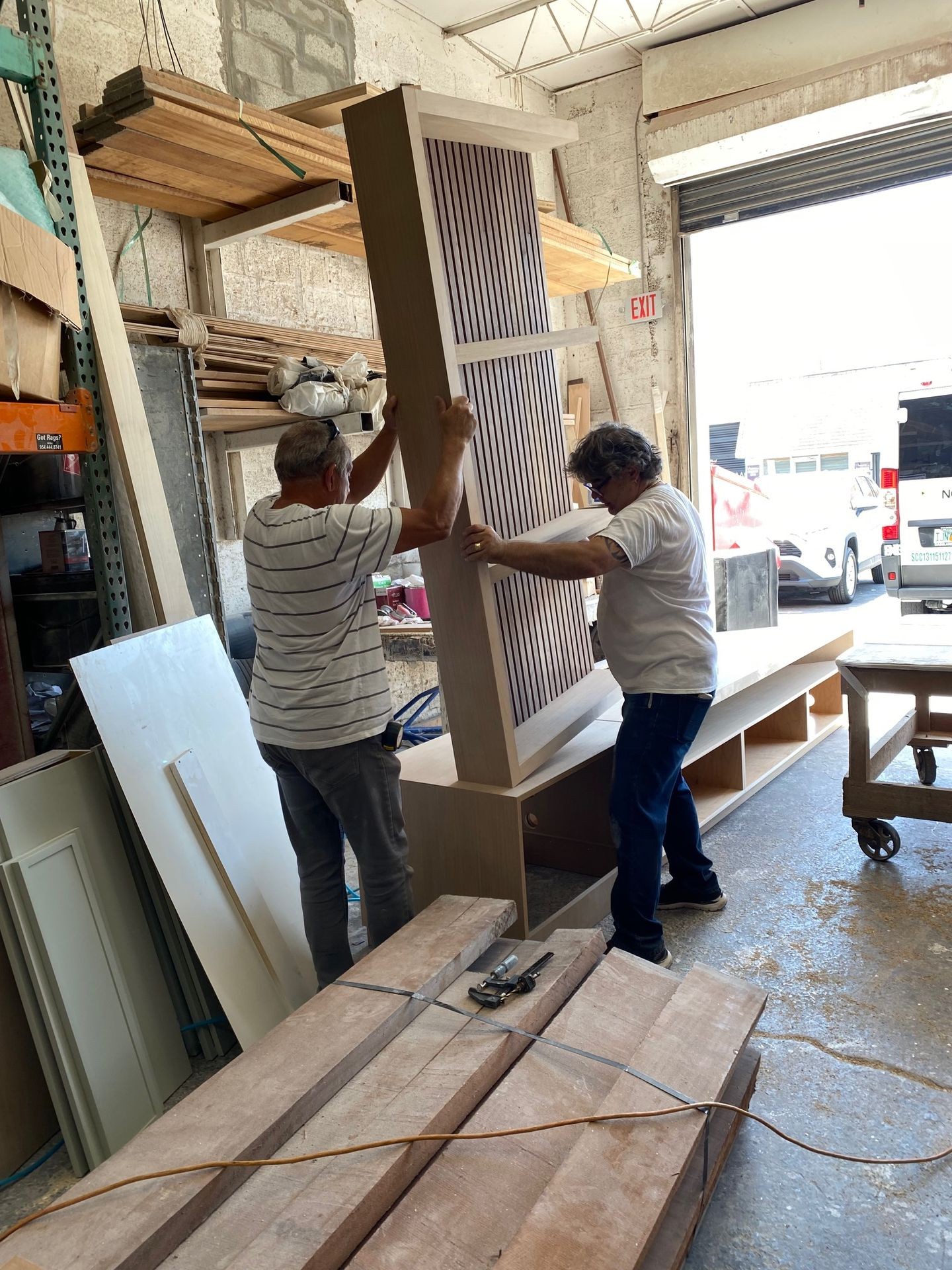 Two people lifting a large wooden piece in a workshop with various tools and materials around them.