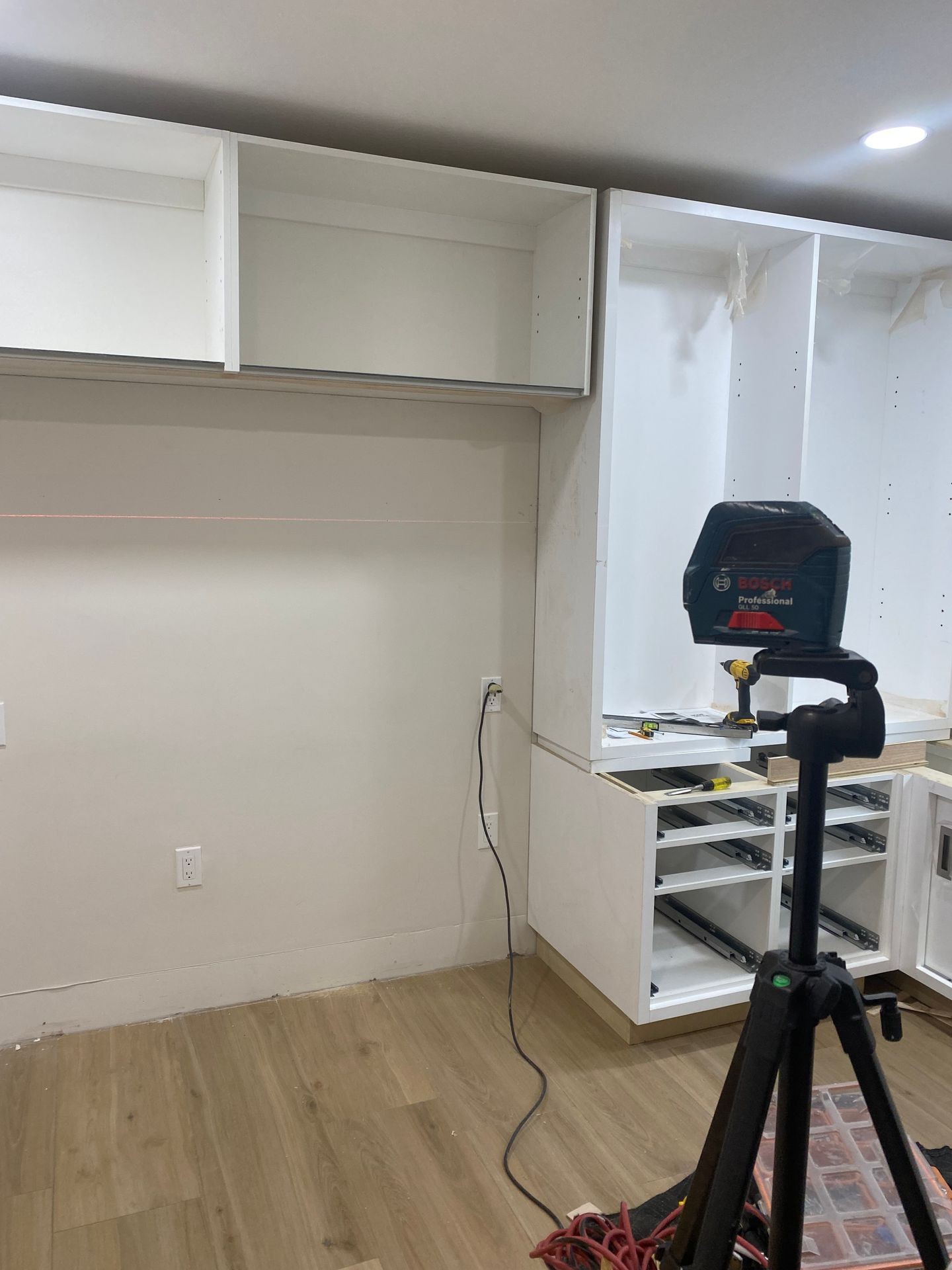 Partially installed white kitchen cabinets with a Bosch professional laser level on a tripod in the foreground.